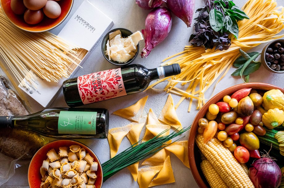 Still-life photography showing dry pasta noodles on a gray background, surrounded by fresh vegetables, eggs, and bottles of wine