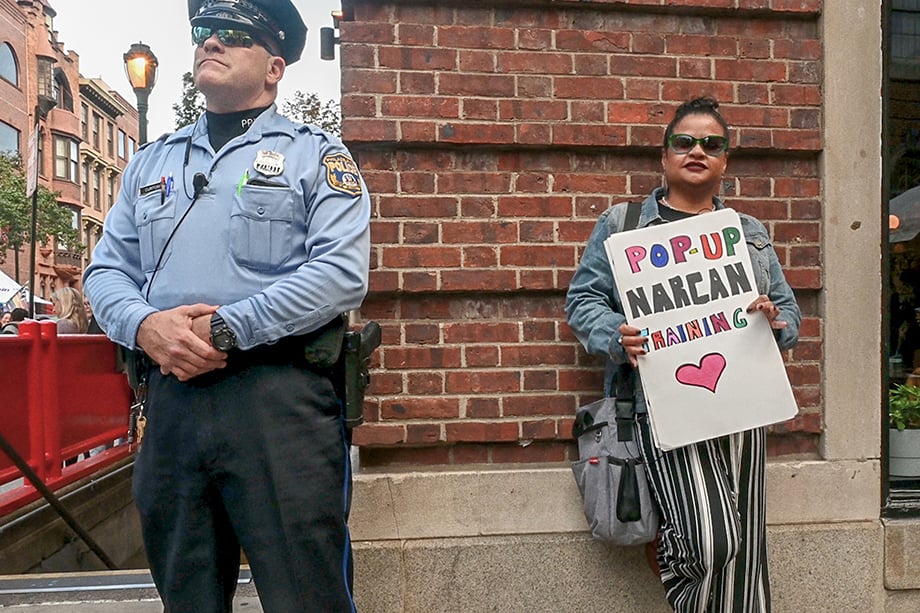 Roz Pichardo stands next to a police officer while holding up a sign that says "pop up narcan training." Photo by Joe Quint. 
