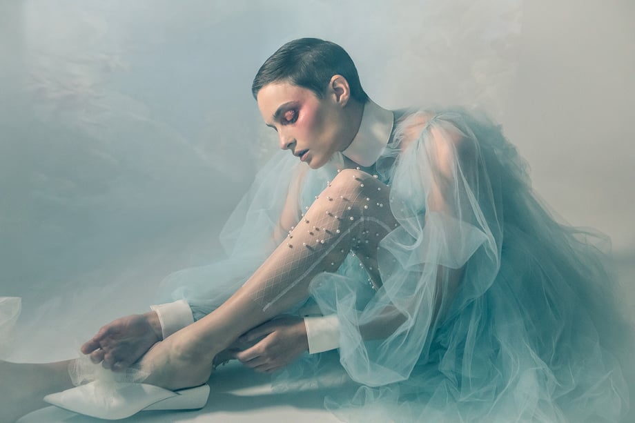 Model Amelia Pool sitting down wearing pearl studded tights and blue toile dress amid a smoky set shot by Kirsten Miccoli