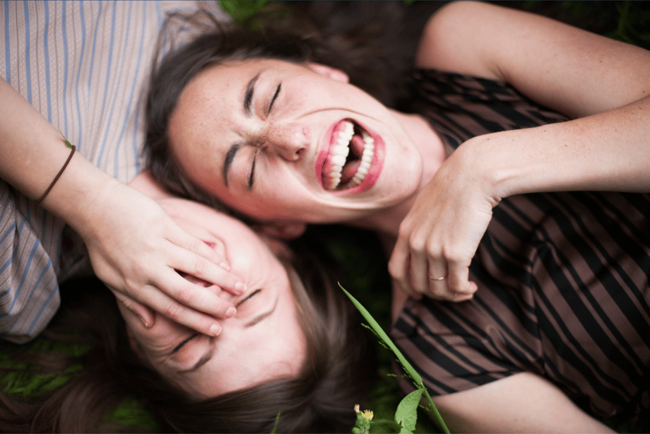 Two young women laying head to head in the grass laughing photographed by Margo Moritz