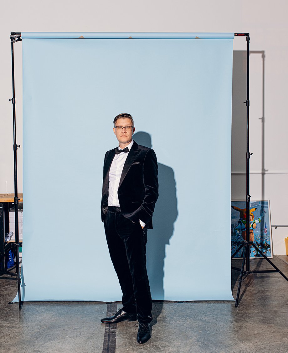 Beeple standing with his hands in his pockets in front of a light blue backdrop while wearing a tuxedo shot by Will Crooks for British GQ