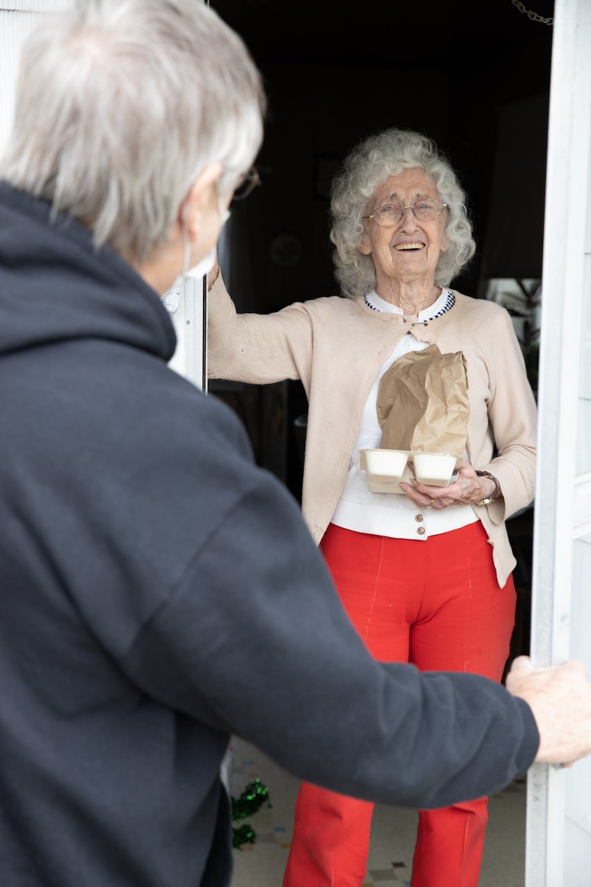 Photo of a Meals on Wheels recipient smiling through her doorway as she receives her food.