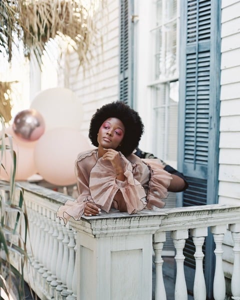 Thi image, taken by Tim for Flaunt Magazine and Badass Balloons, features a young black woman with an afro and bright pink makeup posing on a New Orleans balcony. She is wearing a romantic, frilly sleeved, pink chiffon dress and there are big balloons in the background to match.
