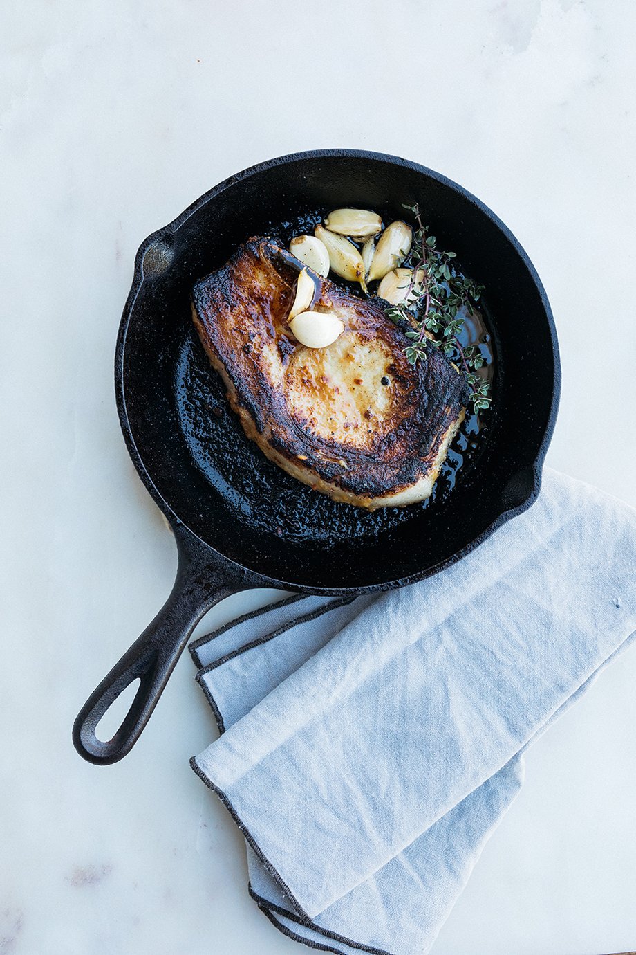 Cast iron pan featuring a seared pork chop and garlic with herbs shot by Mikaela Hamilton for the Women's Heritage Sourcebook