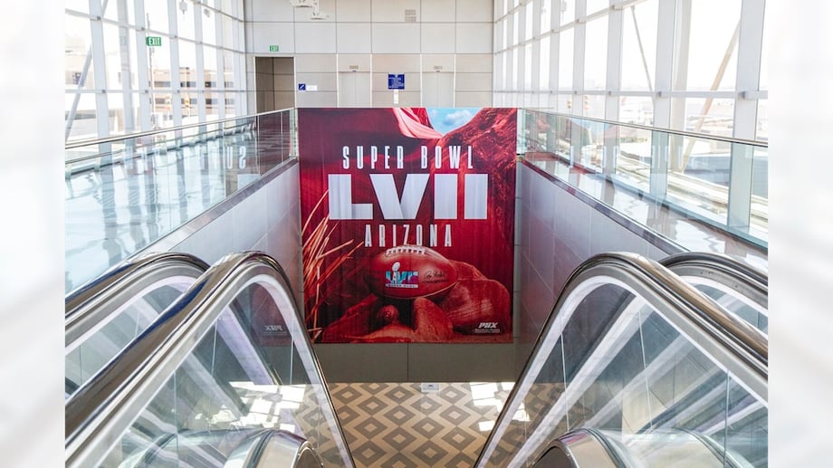 OOH tear sheet of NFL Super Bowl ad displayed over escalator in interior of windowed modern building.