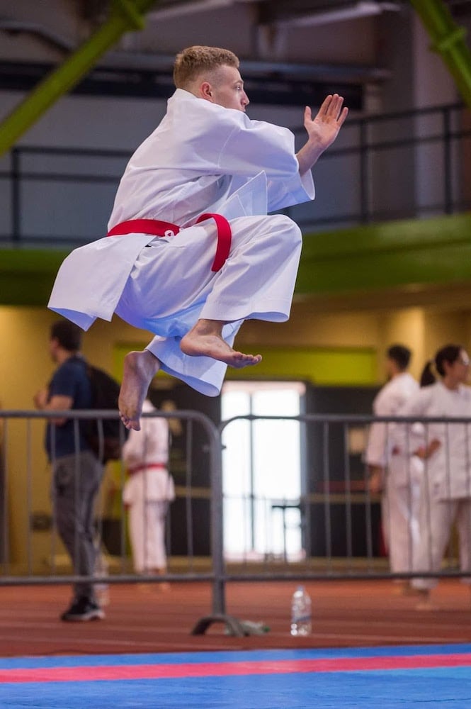 Side profile of karate champion Mihael Ećimović in uniform midair while preforming floor routine at competition.