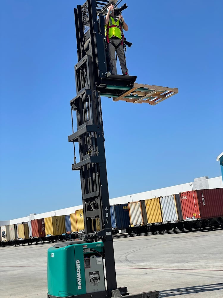 Behind the scenes shot of photographer in safety harness standing on high jack lift with camera, outside the warehouse.