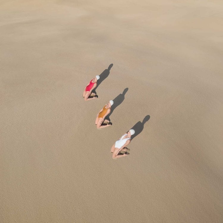 Photo by Brad Walls of three swimmers sitting in the sand casting shadows.