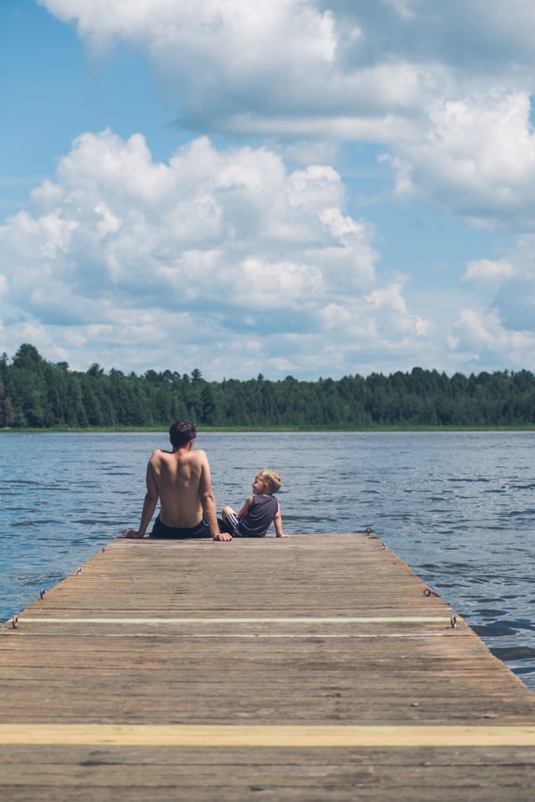 A father and son sitting on the end of a dock, facing a woodsy lake, by photographer CJ Foeckler 
