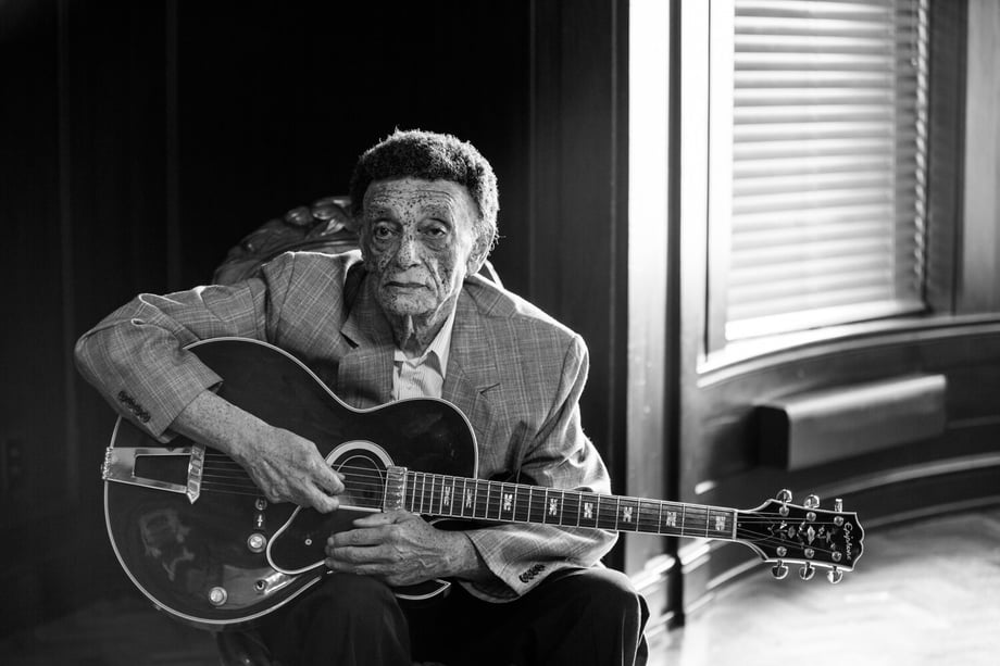 Photo by David Szymanski of an older musician sitting and holding his guitar.