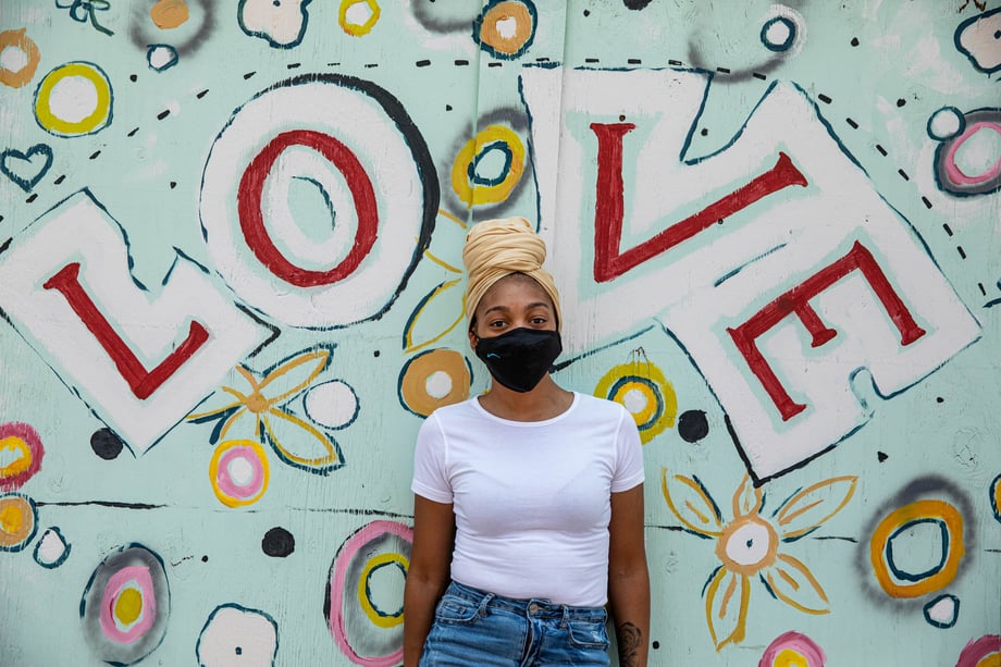 A woman wearing a face mask and head wrap is shown in front of a mural that says LOVE in this photo by Lou Bopp 