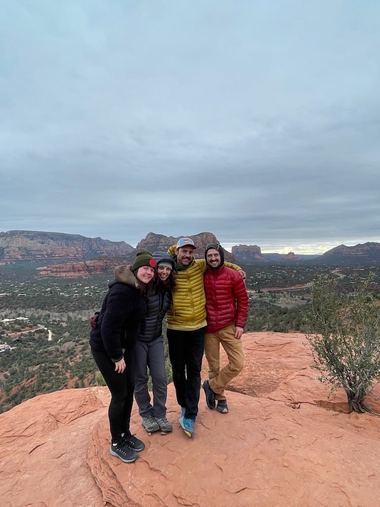 Group photo of four figures standing arm in arm before a desert vista landscape on cloudy day.