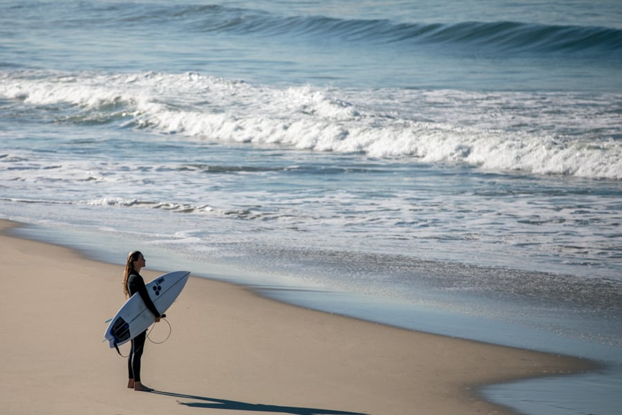 A woman looks out into the ocean, surfboard in hand, by photographer Lou Bopp