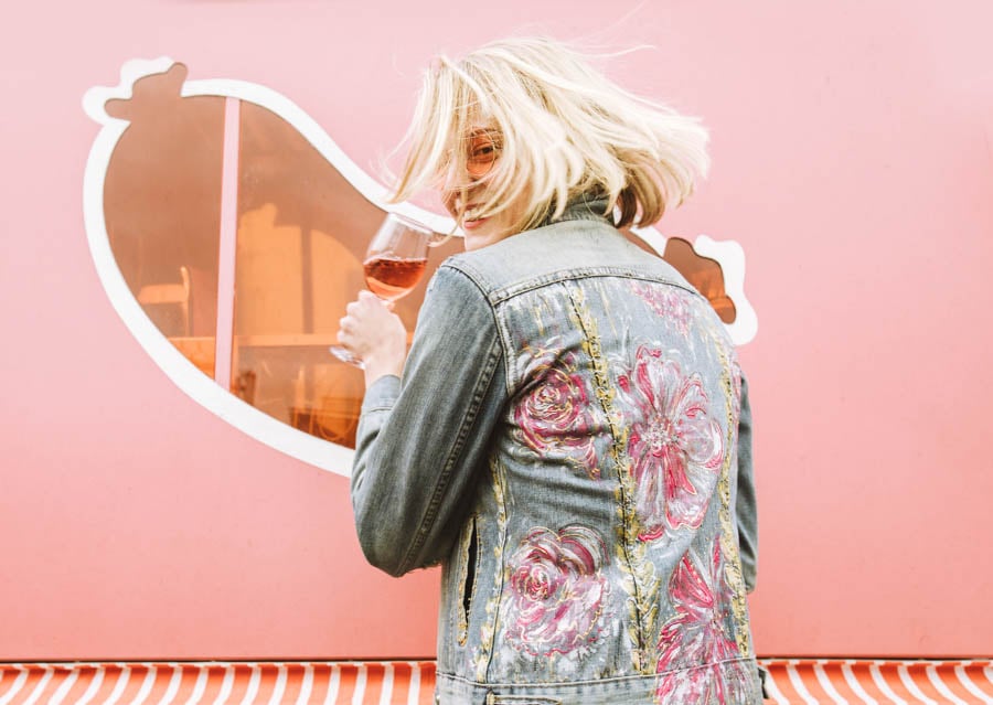 A woman in a pink decored denim jacket smiles in front of a pink wall as she drinks rose by photographer Michelle McSwain of Culver City, Florida.