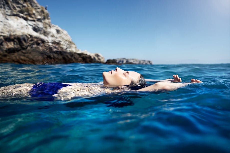A woman in a blue swimsuit floats on her back in blue water next to a steep rock face