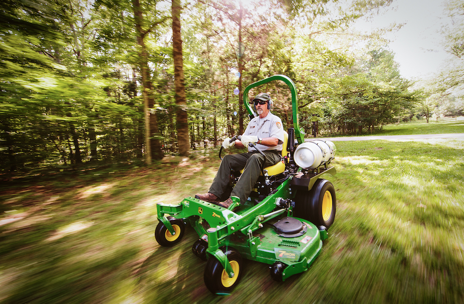  A grassy landscape blurred in the distance, with focus on a man driving a lawn mower at speed