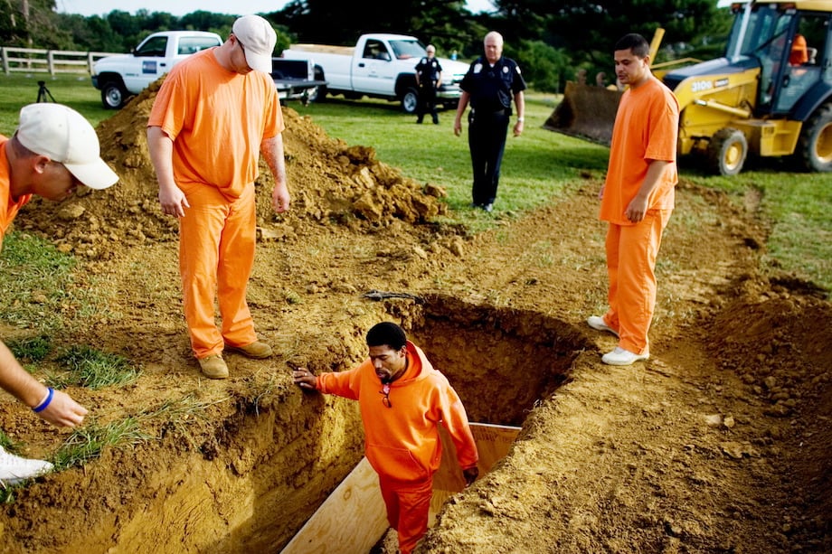 Image of men in orange prison outfits, measuring out and digging a grave while prison wardens oversee them.