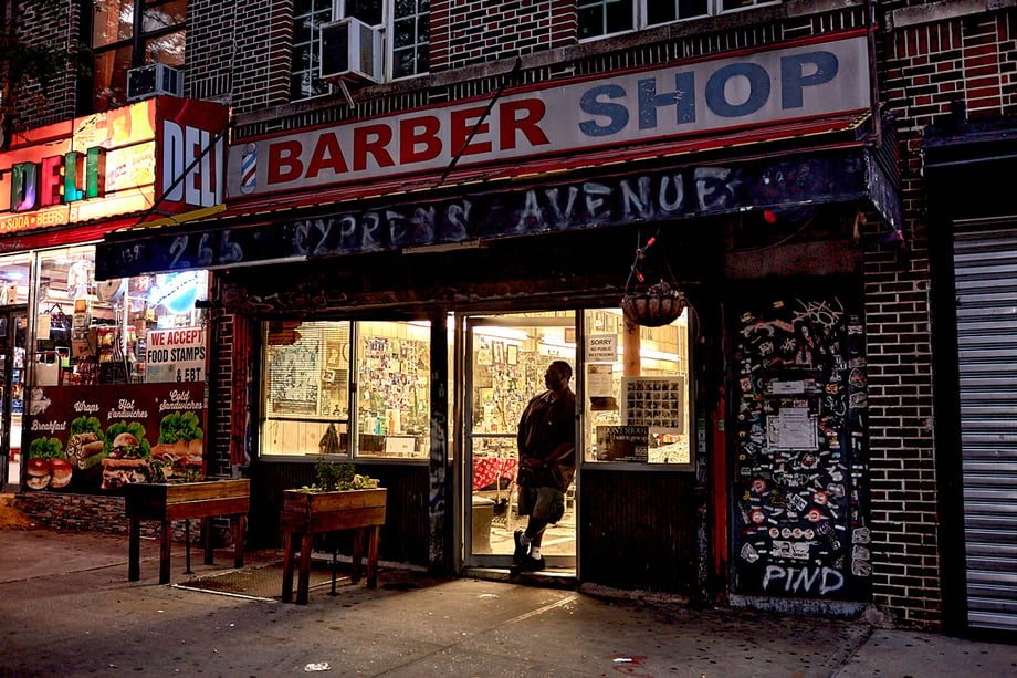 Dusk exterior of a barbershop with a man standing in the open doorway, by Stephen Speranza