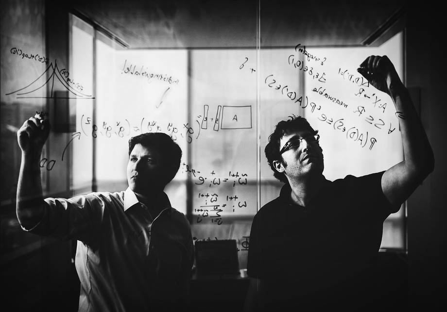 Black and white photo of two men writing math equations on a glass wall taken by Boston-based corporate photographer Adam Glanzman. 