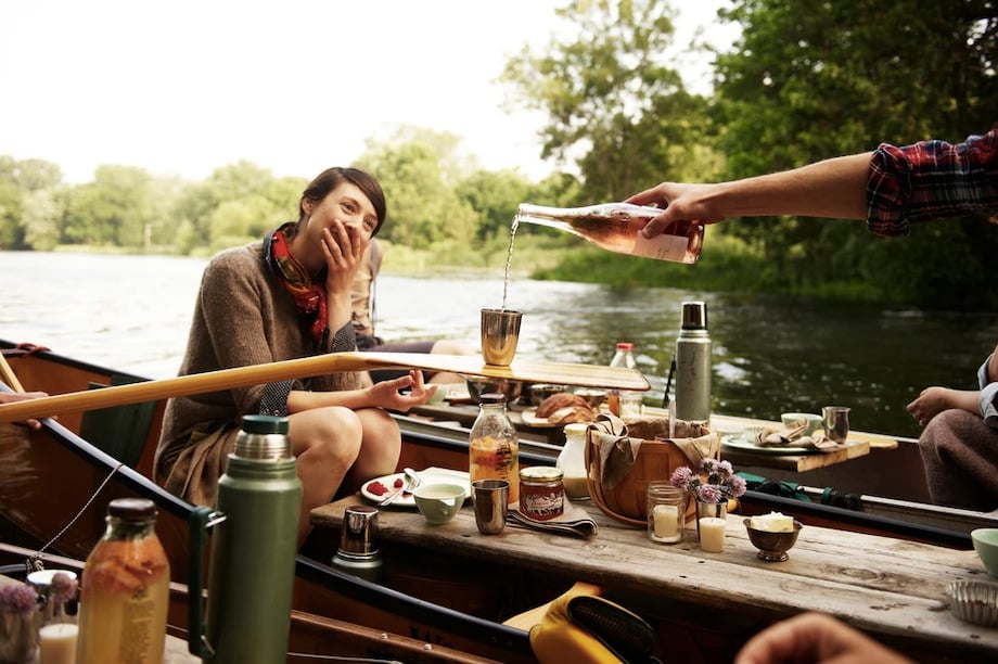 Photo of a lakeside picnic on canoes with figure pouring wine into a cup balanced on a canoe oar, by Atlanta food photographer Ashley Camper.