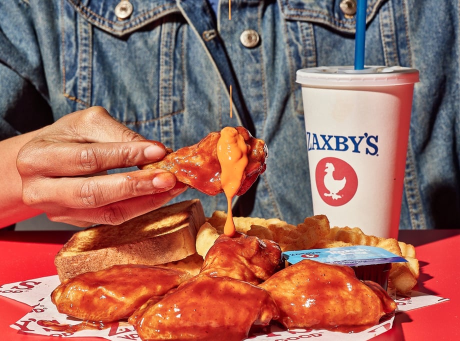 Photo of a hand holding a Zaxby's chicken leg with buffalo sauce, and a Zaxby's cup in the back. 