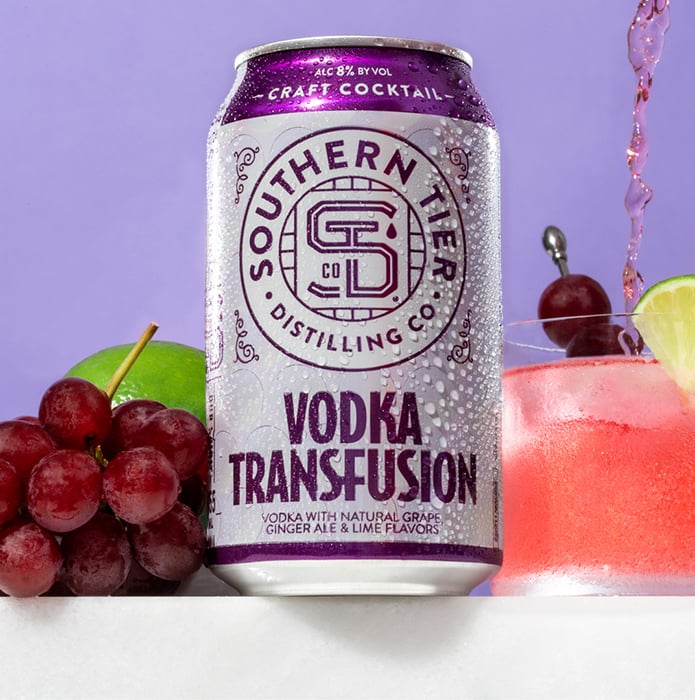 A colorful still life photograph the Vodka Transfusion Canned Cocktail next to fruit, alongside a glass of the pink drink, mid-pour