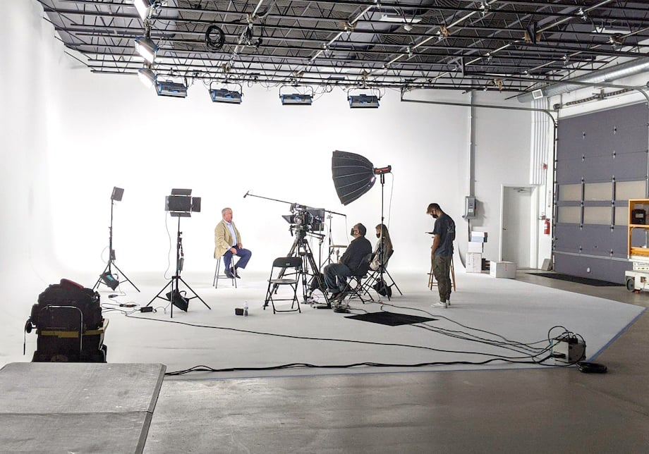 An image of a shoot set-up on a sound stage. You can see the lights, camera, and mic aimed towards the subject who is sitting on a stool with a white cyclorama background.