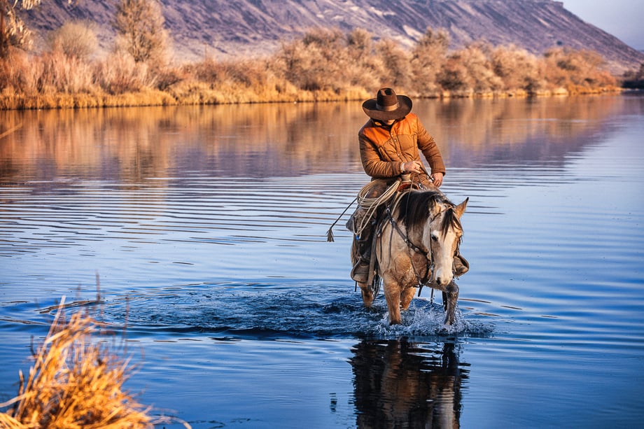 A cowboy riding is horse through shallow waters. 