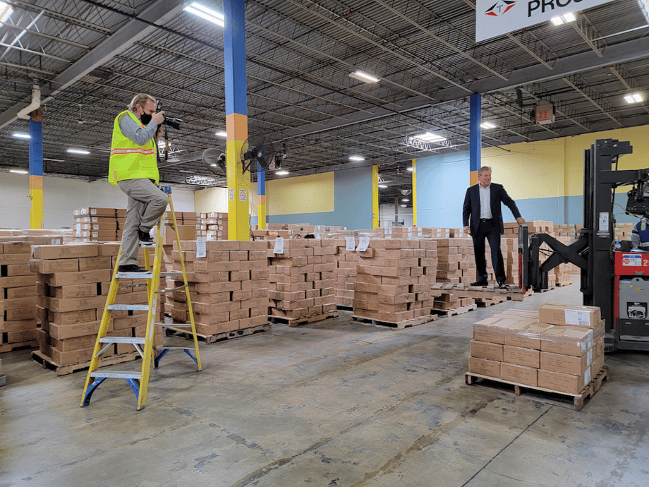 Behind the scenes image of photographer standing on yellow ladder, looking through camera at figure in black suit being lifted on a pallet by a forklift.