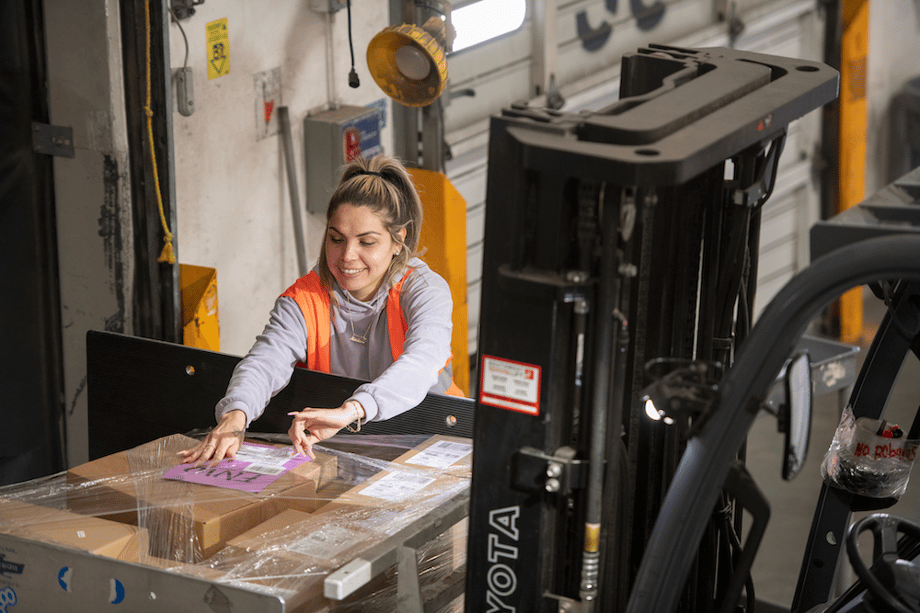 Portrait of figure in orange reflective vest placing label on plastic wrapped skid of boxes, by San Diego-based industrial photographer Frank Rogozienski
