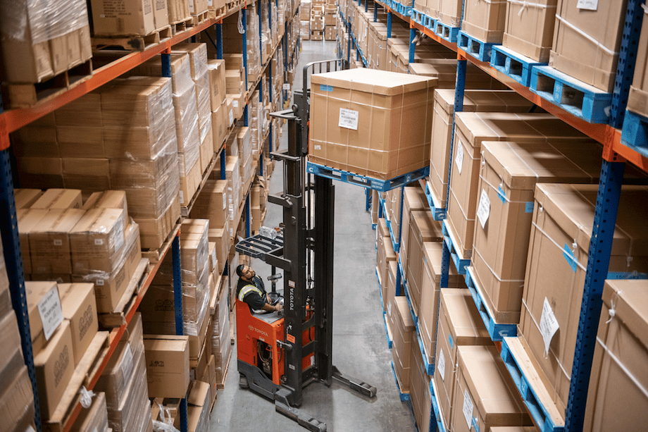 Warehouse photo of figure using black and orange jack lift to unload box from high industrial shelf, by San Diego-based industrial photographer Frank Rogozienski.