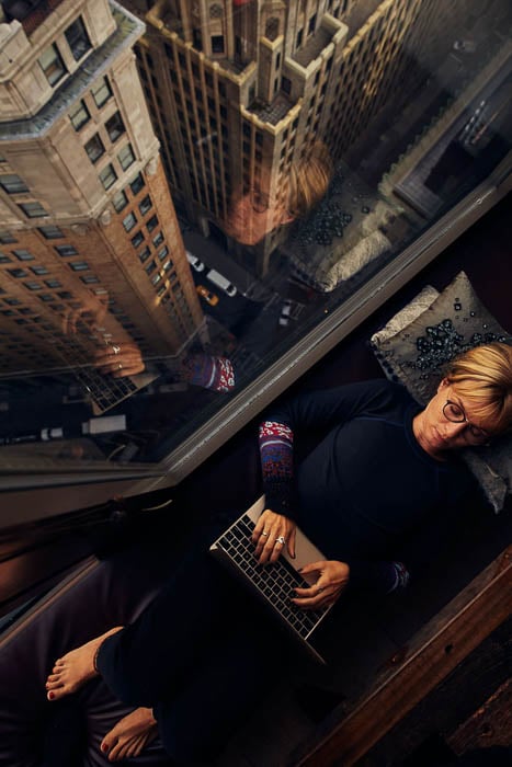 Photo of a woman working on her MacBook beside the apartment window while reclining, taken by New York-based lifestyle photographer Julian Walter