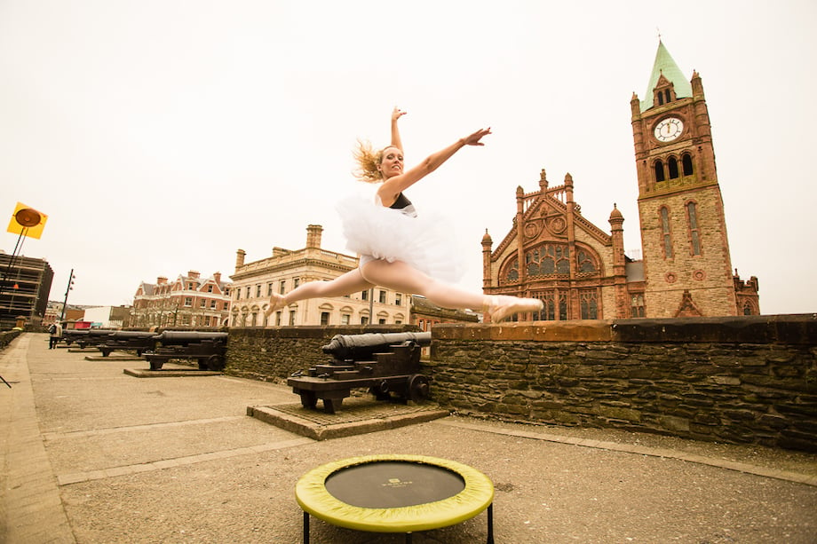 Portrait of a ballerina mid-jump with a trampoline in the foreground and the Guild Hall in the background.