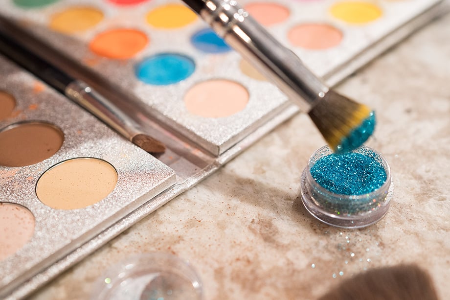 A still life image showing a makeup brush dangling above a container of blue glitter