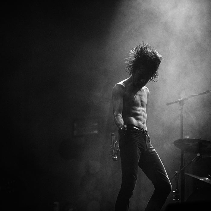 Black and white image of shirtless male musician on stage holding out microphone and dancing shot by Lemar Arecenaux.