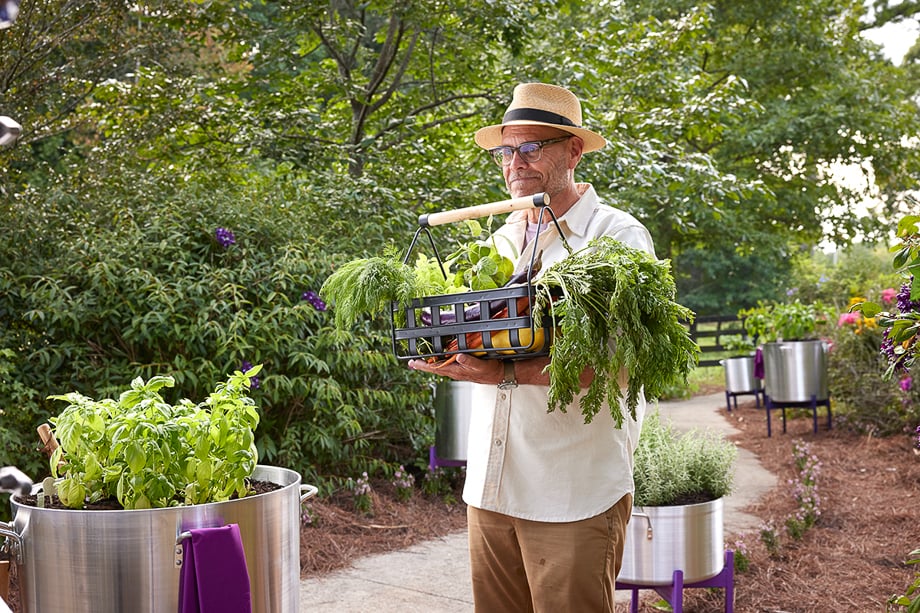 Photo of Alton Brown carrying a container of fresh vegetables at a garden in Serenbe. 