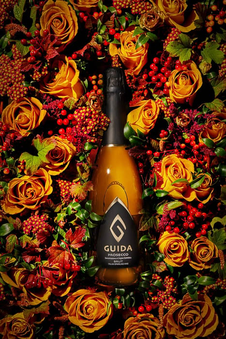 Photo of a Guida Prosecco bottle sitting on a bed of fruit and flowers taken by Miami-based product photographer Marcel Boldú 