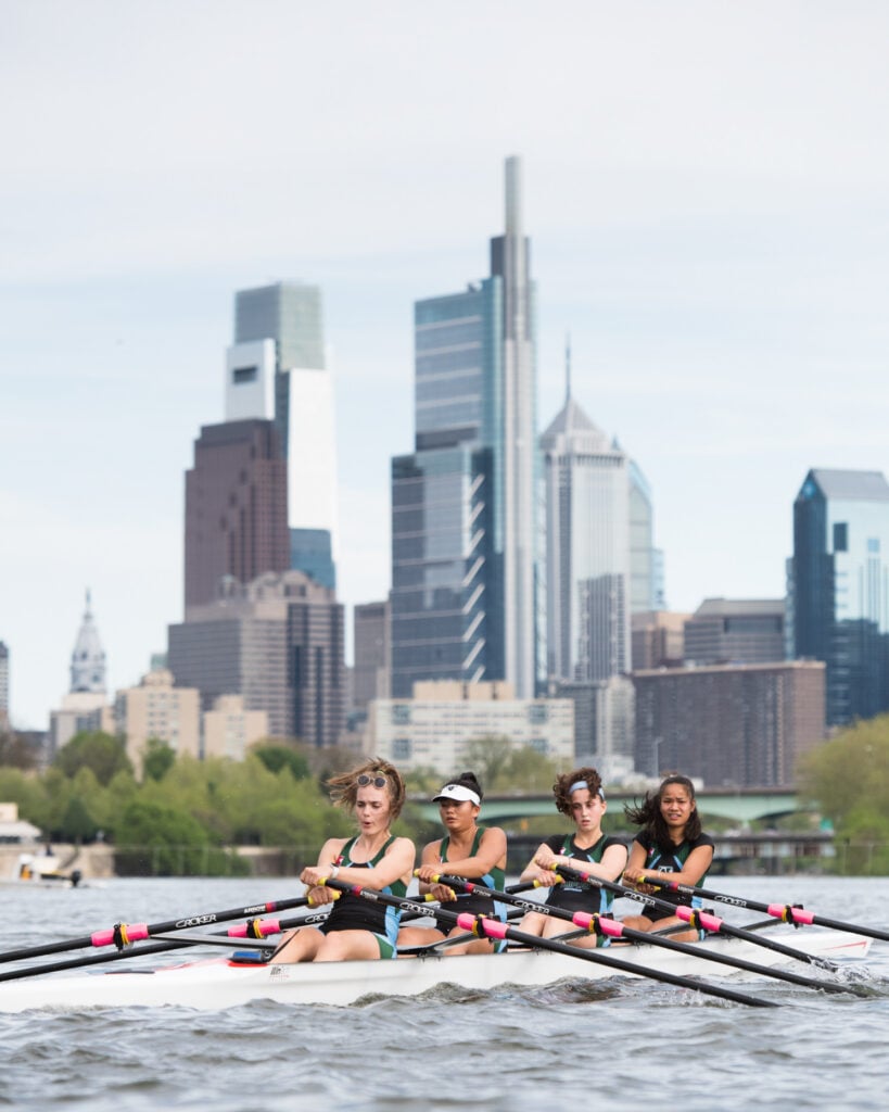Photo by Mark Tassoni of four women on a crew team rowing in the river in front of Philadelphia's skyscapers.
