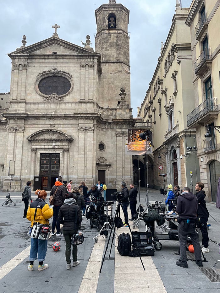 behind the scenes image of the crew and the set-up in a piazza in Barcelona. There are beautiful historical building surrounding the square and the crew have set-up lights and cameras.