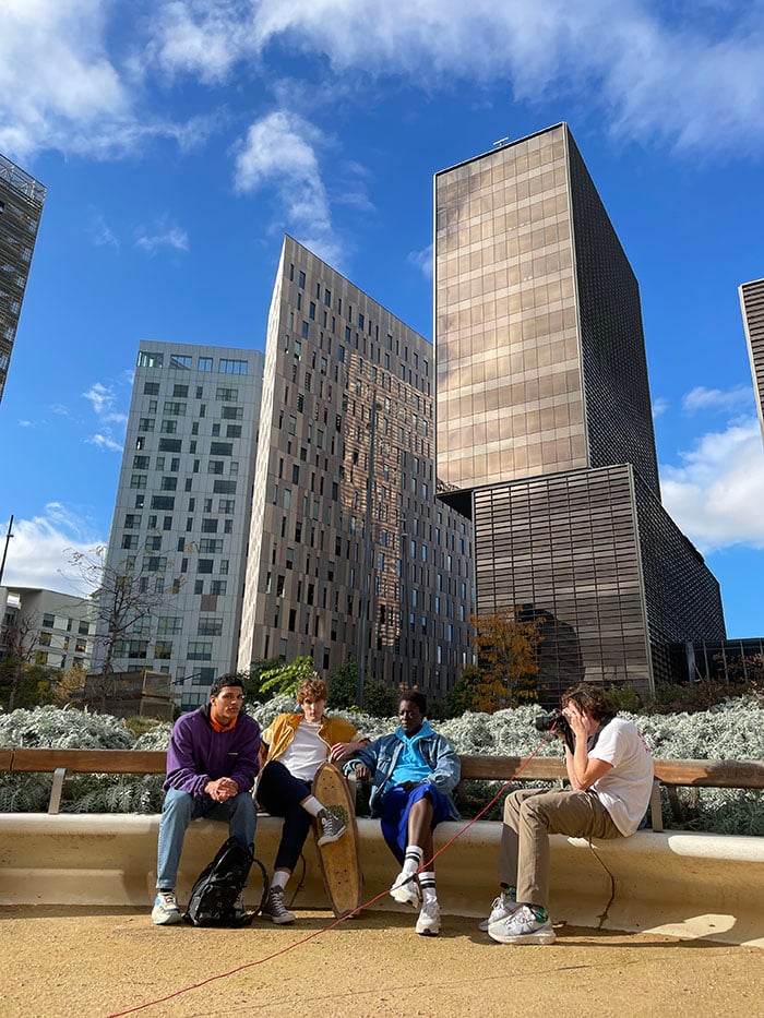 A behind the scenes Image of the photographer taking a shot of three sportily dressed young men who are sitting on a bench. There are big, modern skyscrapers emerging from the park foliage and reaching into the blue sky behind the talent.