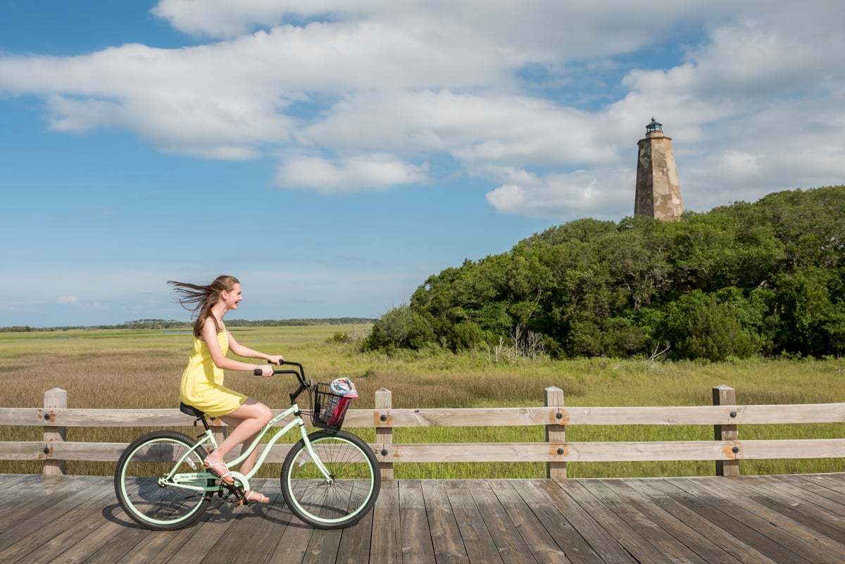 A photo by C2 Photography of a woman in a yellow dress riding a bike on a boardwalk in Bald Head Island.