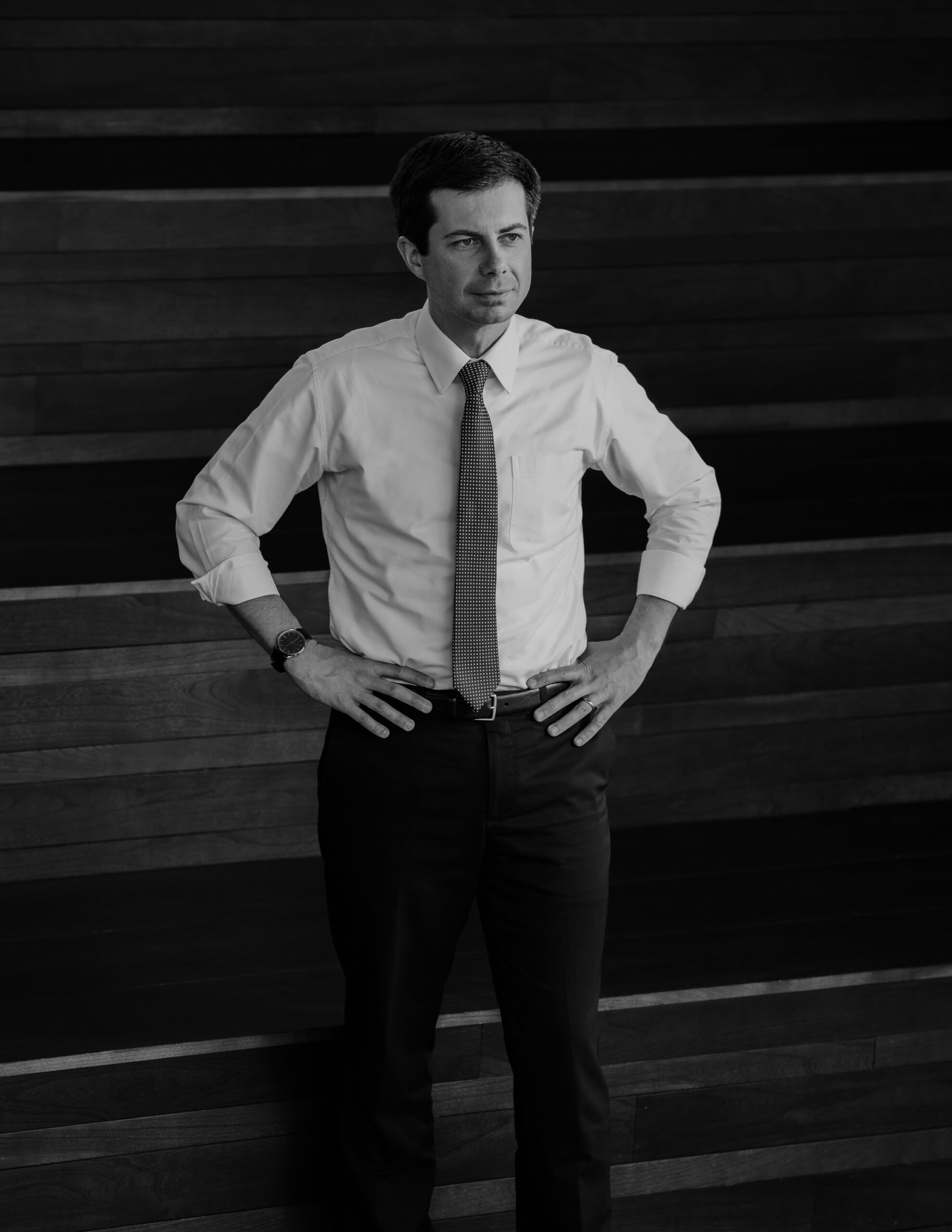 A black and white photograph of Mayor Pete Buttigieg standing with hands on hips looking out of frame