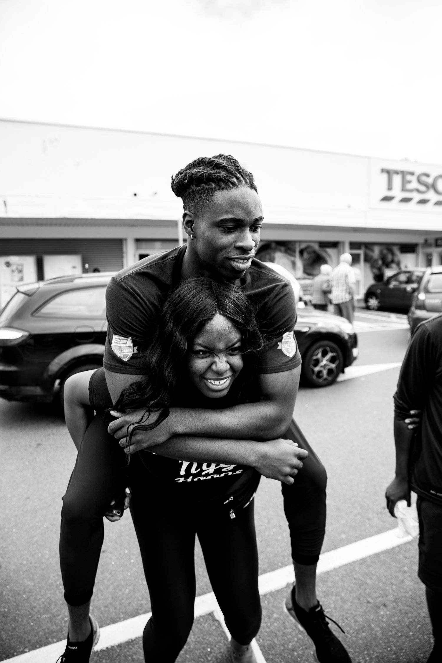 Aidan's cousin carries him on her back in the parking lot of a Tesco in Johnnie Izquierdo's black and white photo