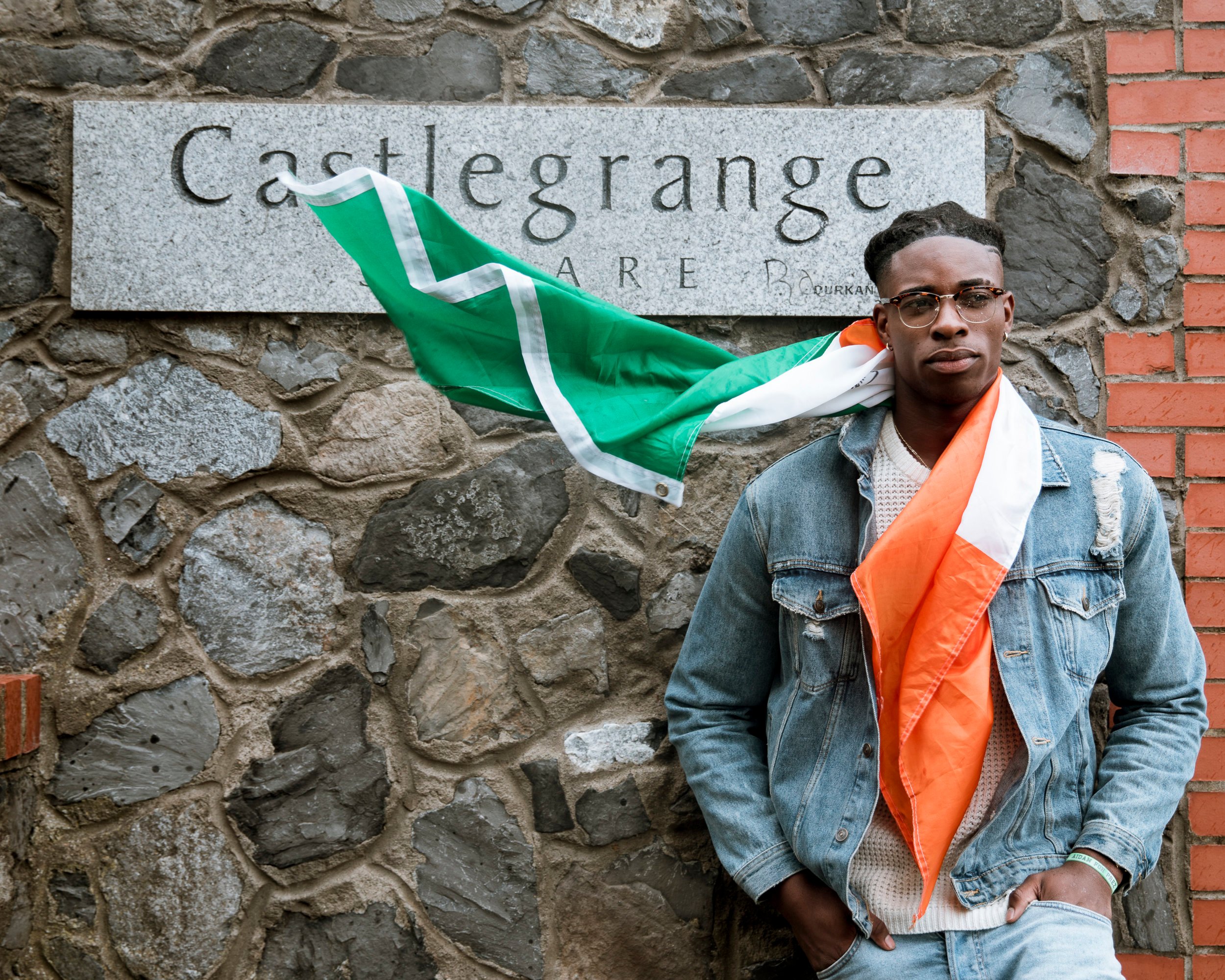 Johnnie Izquierdo stands with the Irish flag draped around his neck in front of a stone wall with a sign reading Castlegrange