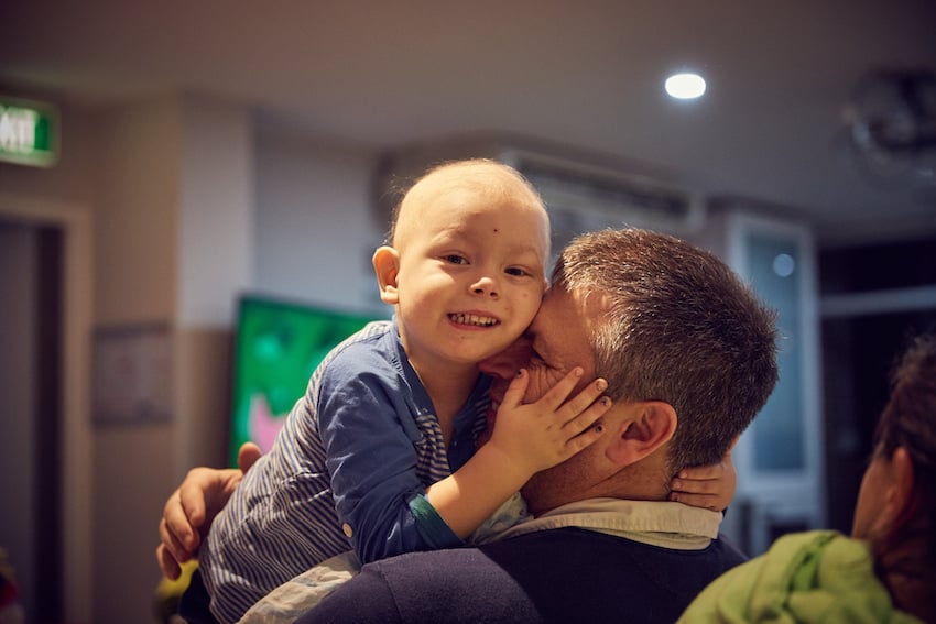 A sick child smiles while embracing their dad at The Ronald McDonald House in Randwick Sydney, by photographer Christian Mushenko. The child is bald, with only minimal wisps of fine, light-colored hair.