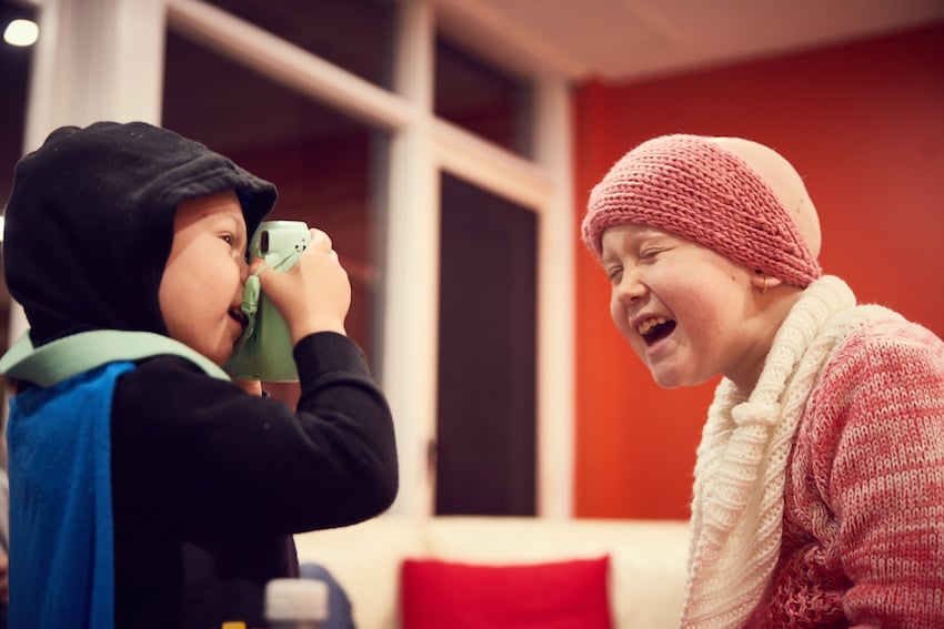 Christian Mushenko's photo of two children playing at The Ronald McDonald House in Sydney. One child is taking a photo of the other with a pastel green polaroid camera. The other child, wearing a pink knitted headband, pink knitted sweater, and white knitted scarf is laughing.