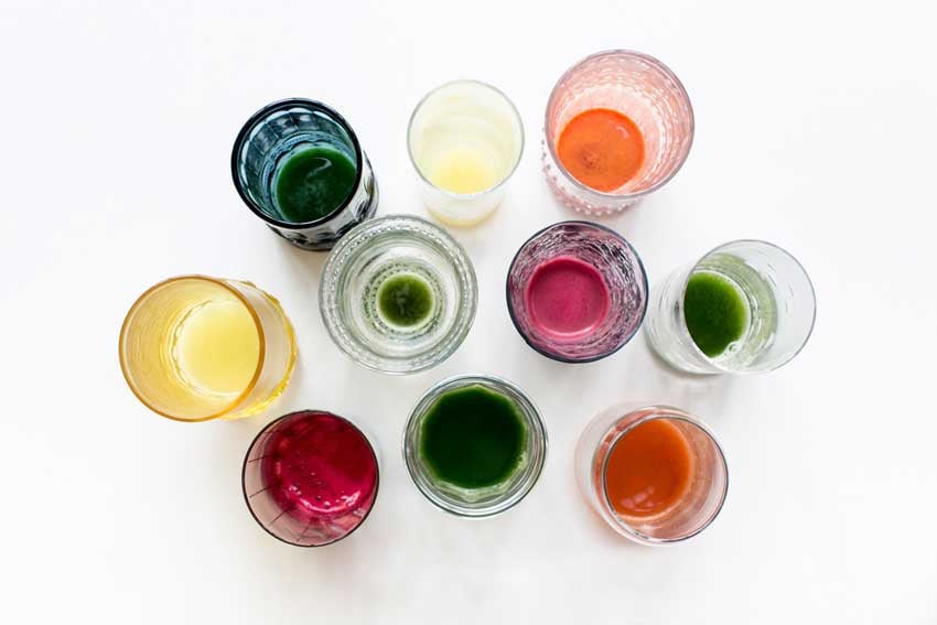 A photo by Brooke Fitts of a selection of different glasses of differently colored juices. The colors of the juices are green, orange, dark pink, and yellow.