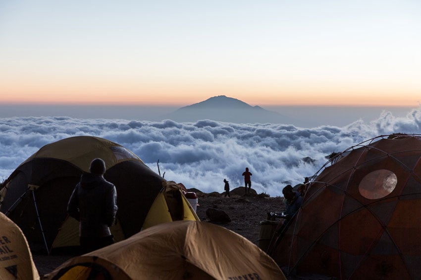 Photographer Clay Cook's image of a mountain peak above the clouds taken from another mountain peak. The photo is taken through a cluster of camping tents and there is a hazy sunrise on the horizon.