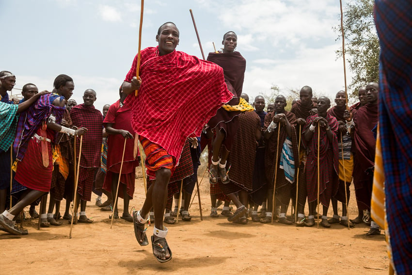 Clay Cook's photograph of an African man in brightly colored tribal clothing smiling and dancing on golden dry earth amidst a circle of others. Everyone is holding a tall stick.