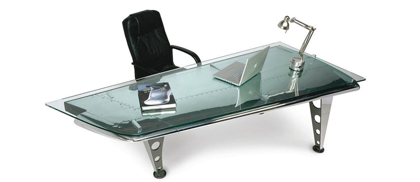 A photo of one of MotoArt's pieces of furniture constructed from an airplane wing. The table has a glass tabletop that sits over a metal segment of an airplane's wing. It has interesting tapering metal legs with gradated holes. The table has a black desk chair at its side for scale and a laptop, metal desk lamp, and a book on its surface.